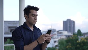 Young Indian man holding smart phone using mobile apps watching funny video. Smiling man having fun chatting in social media. Asian male chatting with friend on Smart Phone white background outdoor.