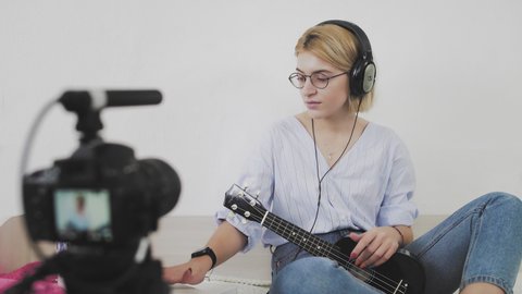 A young woman with glasses is sitting on the sofa, smiling and playing the ukulele to the camera. Playing the ukulele. Video Blogger