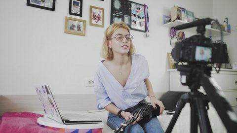 A young woman with glasses is sitting on the sofa, smiling and playing the ukulele to the camera. Playing the ukulele. Video Blogger
