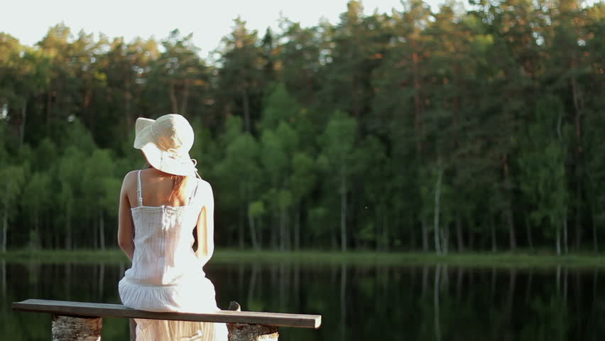 Young beautiful woman in a white dress sits on a wooden pier on the lake, rear view