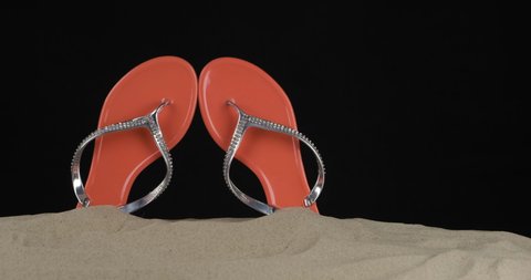 Pair of pink flip flops embellished with rhinestones stick out of the sand. Slider shot. Isolated on black background