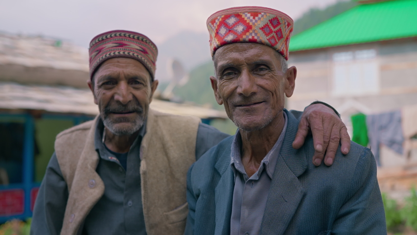 Two happy smiling elderly or old rural Indian male friends or men in traditional attire sitting together looking at the camera in the countryside with village and green mountains in the background | Shutterstock HD Video #1086958082