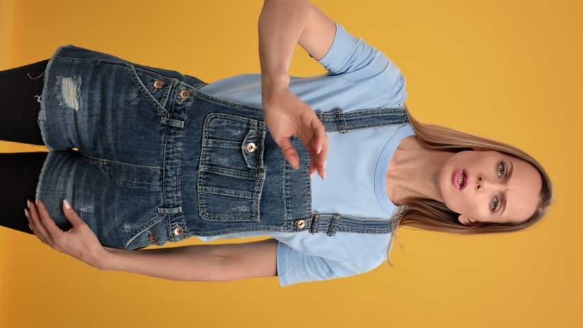 VERTICAL VIDEO POV annoyed modern woman in denim overalls showing gesture no not forbidden stop shaking hand. Portrait of frowning female reject decline disapproval posing isolated on orange Royalty-Free Stock Footage #1086960521