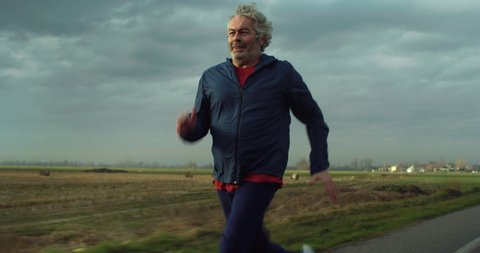 Cinematic shot of motivated athletic retired senior mature male runner in sportswear is running with effort and dedication to maintain himself fit on countryside road surrounded by nature at sunset.