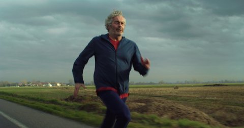 Cinematic shot of motivated athletic retired senior mature male runner in sportswear is running with effort and dedication to maintain himself fit on countryside road surrounded by nature at sunset.