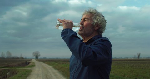 Cinematic shot of thirsty athletic retired senior mature male runner in sportswear drinking water after running to maintain himself hydrated and fit on countryside road surrounded by nature at sunset.