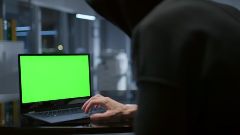 Close up of hacker in hoodie working on laptop with green screen in dark office. Cyber attack and crime concept