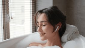 Young Woman Relaxing Taking Bath With Foam Wearing Earbuds Earphones And Listening To Relaxing Music Lying In Bathtub In Bathroom At Home. Spa, Wellness And Relaxation Concept. Side View
