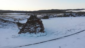 Overflight of Shamanka Rock or Cape Burkhan in winter. View of the village of Khuzhir on the island of Olkhon. Frozen Lake Baikal, covered with snow.