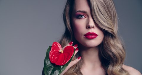 Sexy blonde woman with red lips  holds a red flower. Fashion model  with a long wavy hair posing at studio. Closeup face of a pretty young woman with bright makeup.  Slow motion.