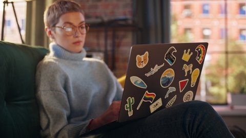 Stylish Female Using Laptop Computer with Diverse LGBT and Lifestyle Stickers on the Back. Young Creative Woman Sitting on a Couch, Typing, Browsing Internet and Checking Social Media at Home.