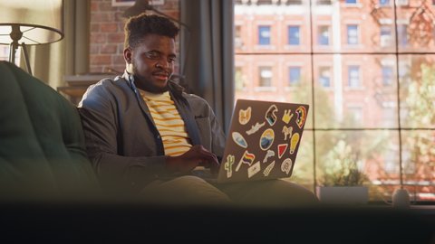 Handsome African American Man Working from Home on Laptop Computer in Sunny Stylish Loft Office. Creative Male Checking Social Media and Browsing Internet on a Couch. Urban City View from Big Window.