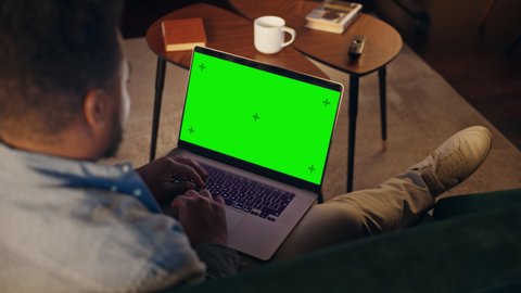 Young African American Man Working from Home on Desktop Computer with Green Screen Mock Up Display. Creative Male Sitting on Sofa, Checking Social Media, Browsing Internet.