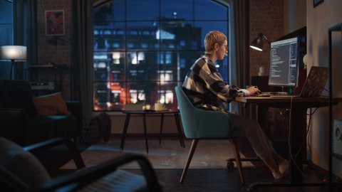 Young Woman Writing Code on Desktop Computer in Stylish Loft Apartment in the Evening. Creative Female Wearing Cozy Clothes, Working from Home on Software Development. Urban City View from Big Window.