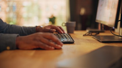 Close Up Footage of Person's Black Hands Use Computer while Sitting at a Table. Creative Designer in a Grey Shirt Using Mouse, Typing on Keyboard. Monitor Display and Laptop on a Desk.