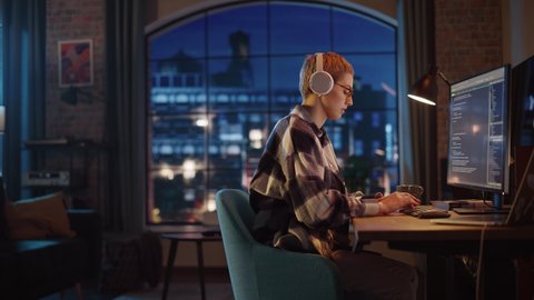 Young Woman Writing Code on Desktop Computer in Stylish Loft Apartment in the Evening. Creative Female Wearing Headphones, Working from Home on Software Development. Urban City View from Big Window.