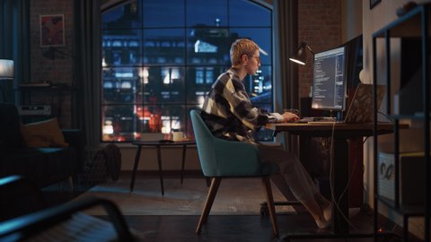 Young Woman Writing Code on Desktop Computer in Stylish Loft Apartment in the Evening. Creative Female Wearing Cozy Clothes, Working from Home on Software Development. Urban City View from Big Window.
