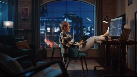 Young Woman Writing Code on Computer in Stylish Loft Apartment in the Evening. Creative Female Wearing Cozy Clothes, Resting Feet on Table and Using Wireless Keyboard. Urban City View from Big Window.