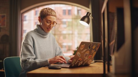 Young Diverse Woman Working from Home on Laptop Computer with Stickers in Sunny Stylish Loft Apartment. Creative Designer Wearing Cozy Blue Sweater and Glasses. Urban City View from Big Window.