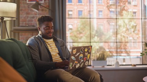 Handsome African American Man Working from Home on Laptop Computer in Sunny Stylish Loft Office. Creative Male Checking Social Media and Browsing Internet on a Sofa. Urban City View from Big Window.