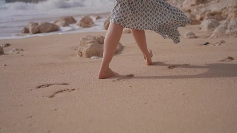 The girl walks barefoot on the sand towards the sea, leaving footprints in the sand. The girl walks away from the camera on the beach. The sea in the background