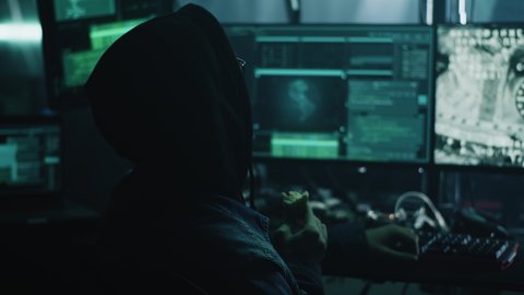 Anonymous hacker in hoodie eating and disrupting space unit coupling process remotely from computers while sitting in dark room of criminal base. Screen footage furnished by NASA.
