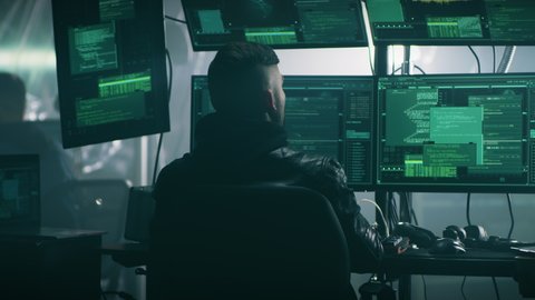 Male hacker raising hands behind head while police in uniform with guns arresting him during a cyber attack in a dark gloomy room with screens