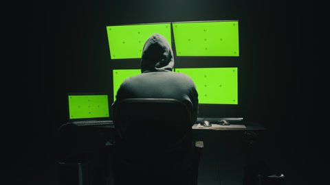 Pan around concept shot of anonymous man in hoodie writing code to hack database while sitting at desk with computer monitors, with green screens chromakey in dim room of hacker base