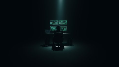 A cybercriminal in an anonymous mask looking at the camera in a dark room with computers during hacking data at night