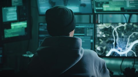 Back view of young man in hoodie and hat typing on computer keyboard and spying on victim while sitting at table in dark room