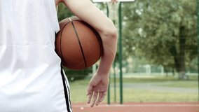 Boy standing on a basketball court and holding a basketball under his arm. Video from the back shot of teenager standing with a ball in his hand and a basketball basket in front of him. 
