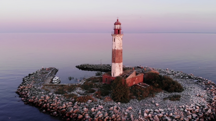 Old lighthouse at small artificial island, aerial shot in dusk, alarmed birds fly around building and sometimes flash near the camera. Despite shabby look of tower and abandoned house near