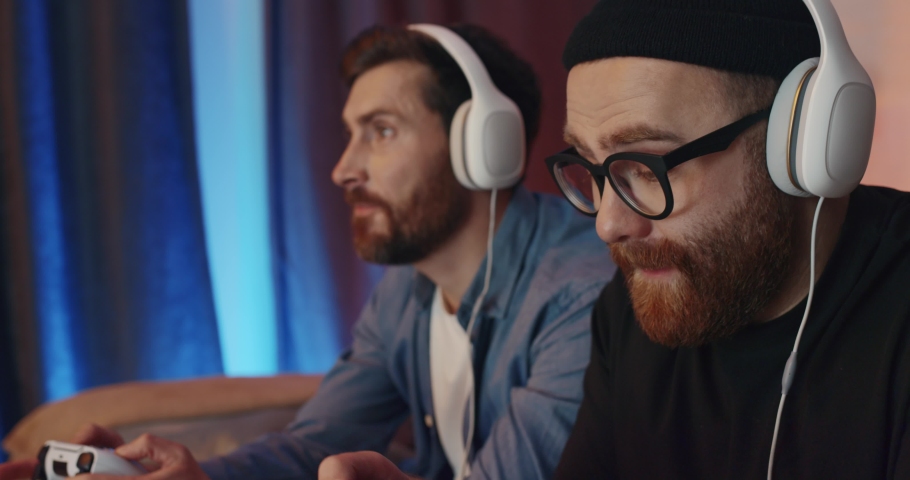 Two Men Friends sitting at Home wears Headphones playing Video Games and Lose. Handsome Males using PlayStation and holding Joystick for Playing Games looking at TV. Technology. Quarantine Leisure. Royalty-Free Stock Footage #1086984302