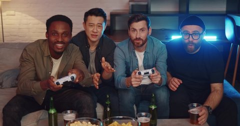 Attractive Men Resting at Home playing Video Games at Home and Losing. Group of Male Friends having a Night Party sitting on the couch with Snacks and Drinks. Playing Computer Games using Controllers
