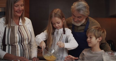 Portrait of Loving Grandmother Teaches a Little Girl to Cook and Beats Eggs. Cute Granddaughter Cooking Dough while Mature Couple Man and Woman with a Little Boy standing near in the Kitchen.