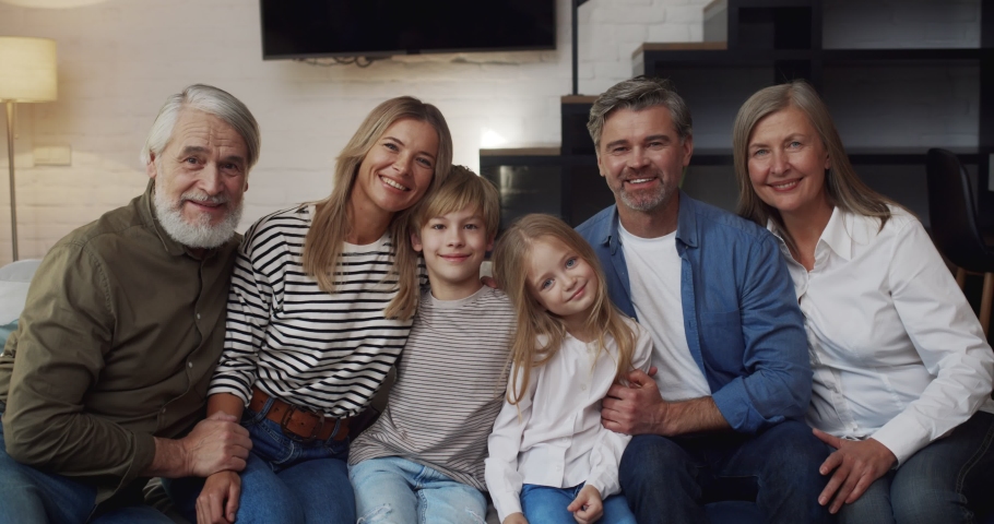 Portrait of Caucasian Big Family sitting on the sofa at Home. Children sitting Between Loving Parents and Grandparents Hugging while looking into the camera.  Generation