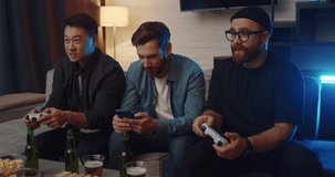 Handsome Men Friends sitting on couch playing Video Games one Man Browsing Phone. Multinational friends using PlayStation for home leisure spending free time. Technology. Gamers. Console. Victory