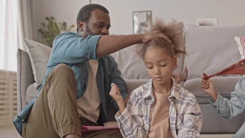 Medium shot of loving African-American father making perfect ponytail to his little daughter sitting on carpet at cozy living room