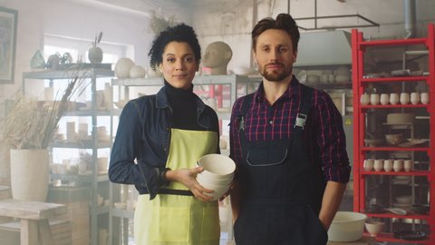 Portrait of couple of young potters, woman and man, standing in workshop and holding ceramic items, looking at camera and smiling, sharing positive emotions, Zoom in, Slow motion.