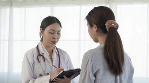 4K, A white Asian female doctor standing and taking notes on a long-haired Asian patient wearing a long-sleeved shirt in a hospital examination room. Concept Health and Medicine