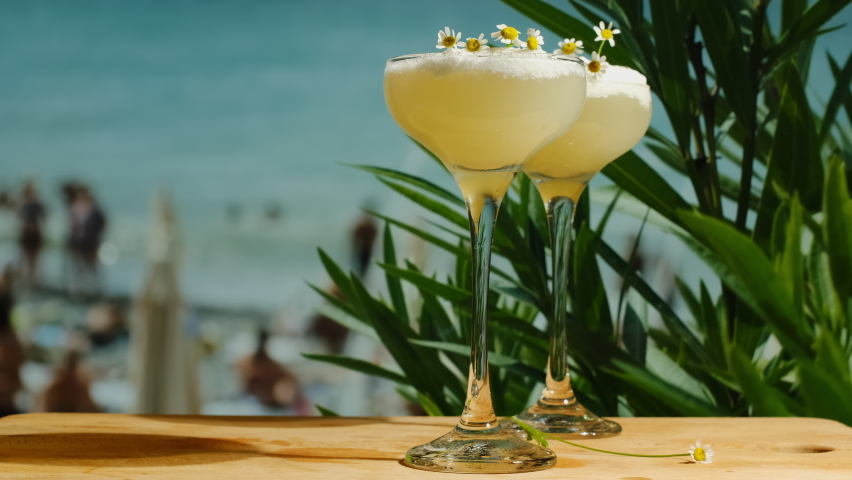 Beach bar. Foamy alcoholic lemon cocktails on table bar, sea on background. Two drinking glasses of pisco sour cocktails decorated fresh chamomiles. Summer sea resort concept.