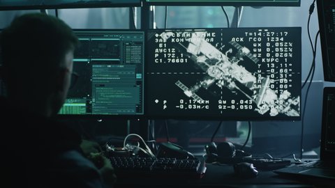 A hacker in glasses at a computer with software, during a cyber attack on the docking of the space module to the international space station in dark refuge. Footage on the screeen furnished by NASA.