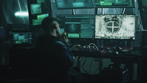 Young man eating donut and hacking process of spaceship and space station coupling docking while sitting at desk in dark room at night. Footage on the screeen furnished by NASA.