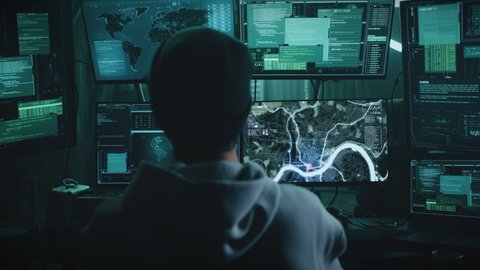 Zoom out view of anonymous man in hoodie and hat using spy software on computer to locate victim, while sitting at desk on dark hacker base