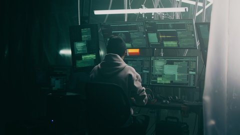 Policemen in armor putting handcuffs on cyber criminal sitting at desk with computers during mission on dark hacker base