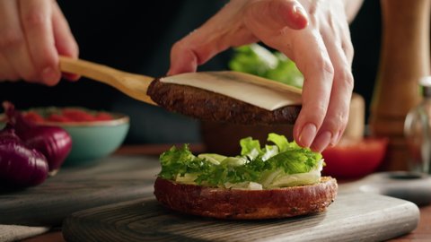 Chef cooking burger, putting meat chop with cheese on lettuce close-up, making sandwich, fast food concept, recipe of preparing homemade hamburger with vegetables.