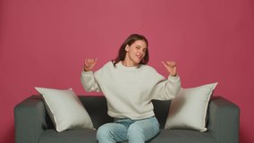 Joyful young girl dancing listen to music, having fun, celebrating win, sitting on couch on pink studio background
