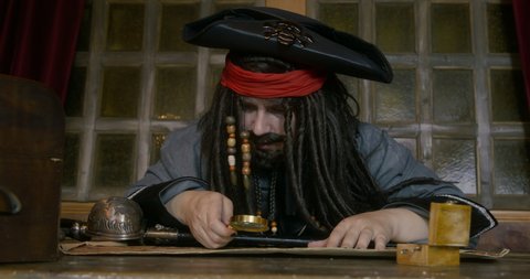 Pirate captain with dark long hair in cocked hat looks through magnifying glass on old sea map AT table searching for treasures in cabin
