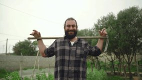 Slow motion video of bearded man holding hoe over its shoulders in garden smiling to camera