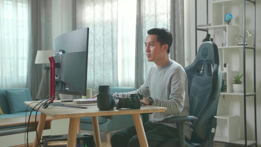 Asian Cameraman In Long Sleeved T-Shirt And Black Pants Looking At Desktop Computer Screen And Writing In Notebook On A Table While Working At Home